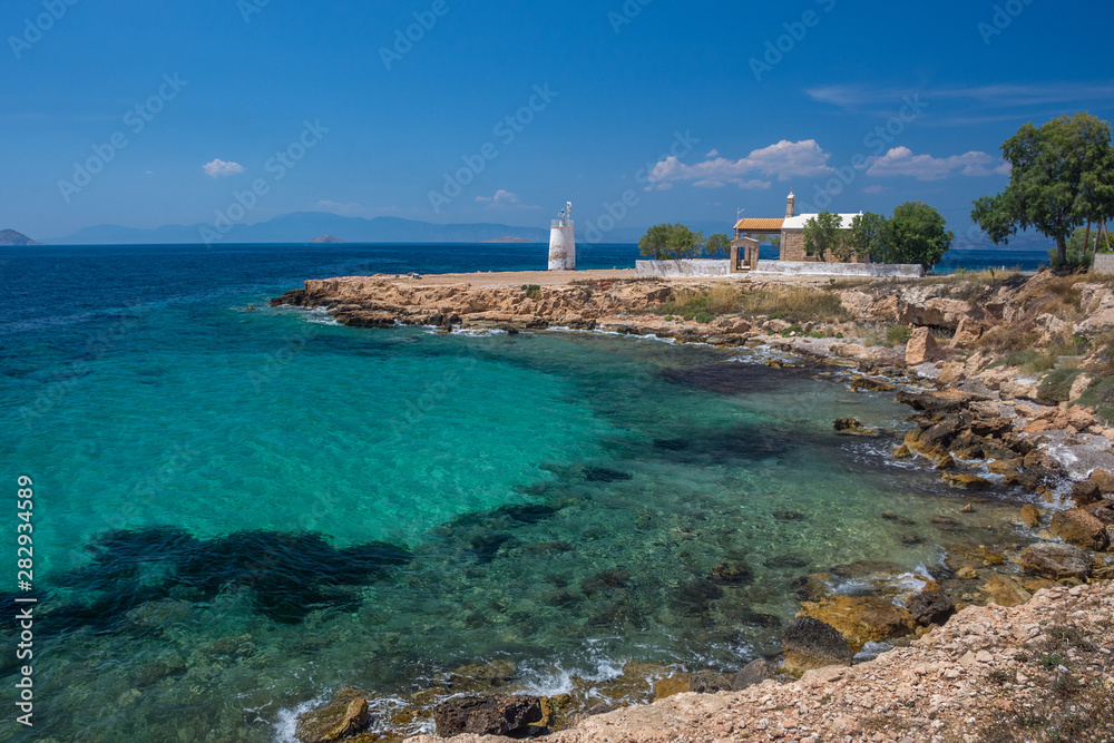 The wild coast of Aegina island with clear and blue waters of Mediterranean sea and the old small lighthouse in the background, in Saronic gulf, Greece.