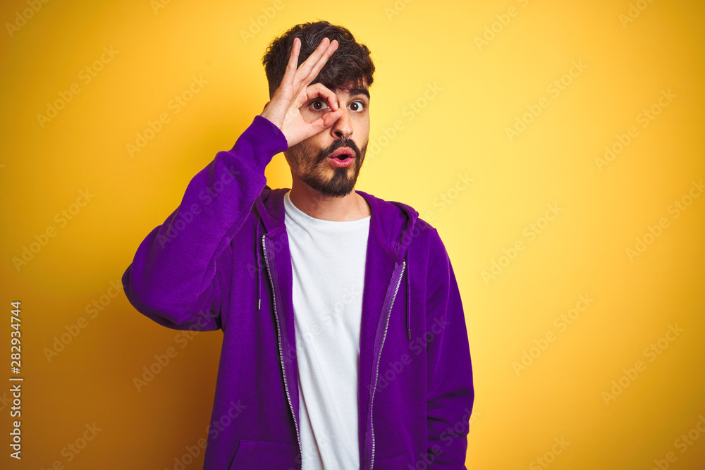 Young man with tattoo wearing sport purple sweatshirt over isolated yellow background doing ok gesture shocked with surprised face, eye looking through fingers. Unbelieving expression. Young man with tattoo wearing sport purple sweatshirt over isolated yellow background doing ok gesture shocked with surprised face, eye looking through fingers. Unbelieving expression.