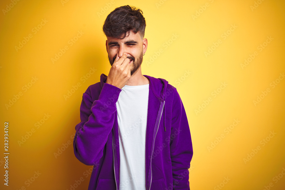 Young man with tattoo wearing sport purple sweatshirt over isolated yellow background smelling something stinky and disgusting, intolerable smell, holding breath with fingers on nose. Bad smells Young man with tattoo wearing sport purple sweatshirt over isolated yellow background smelling something stinky and disgusting, intolerable smell, holding breath with fingers on nose. Bad smells