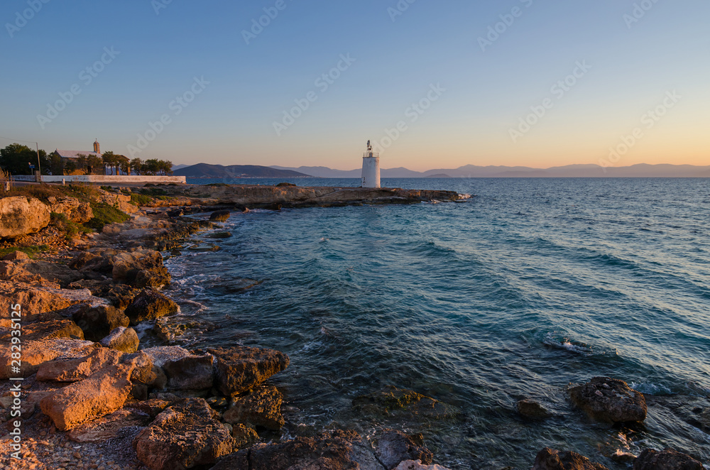 Wild coast of the Aegina island and the old small lighthouse in the background, Saronic gulf, Greece, at sunset.