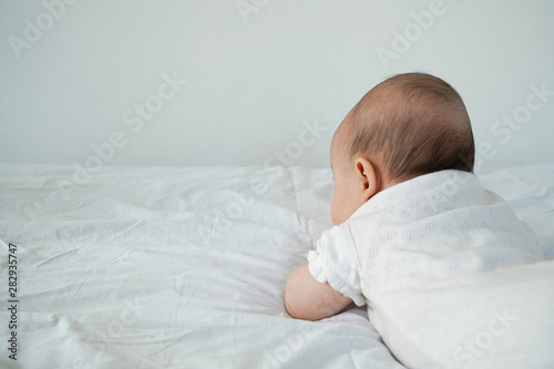 The baby lies on his stomach. The baby looks upward at the wall. The back of the head of a newborn