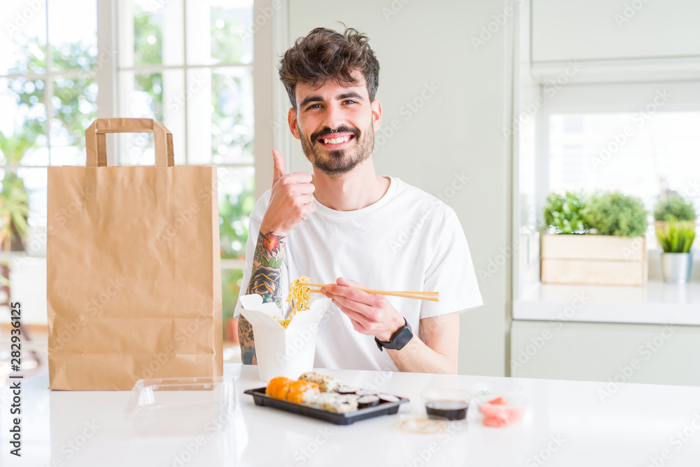 © Krakenimages.com - Young man eating asian sushi from home delivery happy with big smile doing ok sign, thumb up with fingers, excellent sign