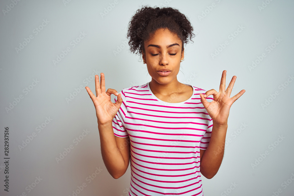 African american woman wearing casual pink striped t-shirt over isolated white background relax and smiling with eyes closed doing meditation gesture with fingers. Yoga concept. African american woman wearing casual pink striped t-shirt over isolated white background relax and smiling with eyes closed doing meditation gesture with fingers. Yoga concept.