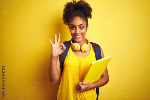 Afro woman using backpack and headphones holding notebook over isolated yellow background doing ok sign with fingers, excellent symbol