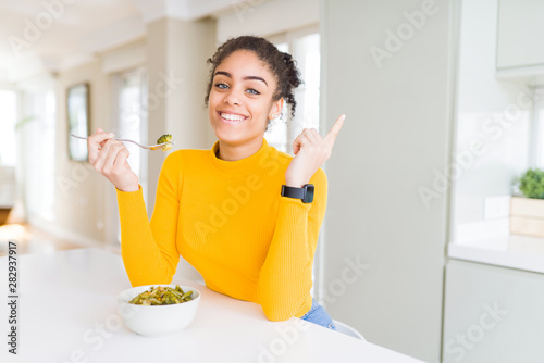 Young african american woman eating healthy green vegatables very happy pointing with hand and finger to the side