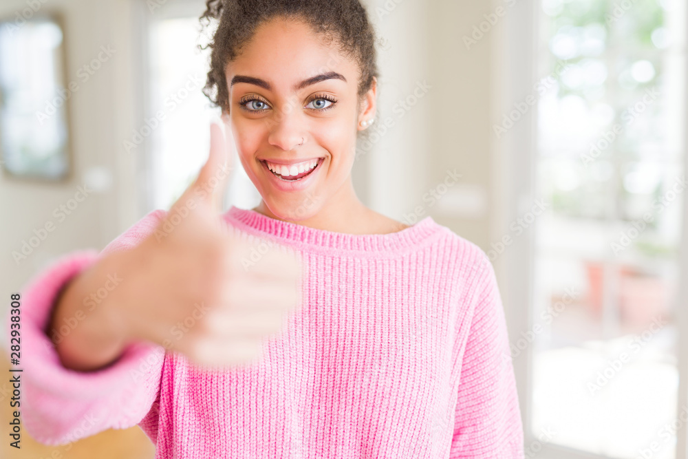 Beautiful young african american woman with afro hair doing happy thumbs up gesture with hand. Approving expression looking at the camera showing success.
