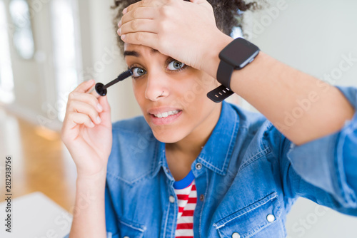 Young african american girl applying eyelashes mascara stressed with hand on head, shocked with shame and surprise face, angry and frustrated. Fear and upset for mistake.