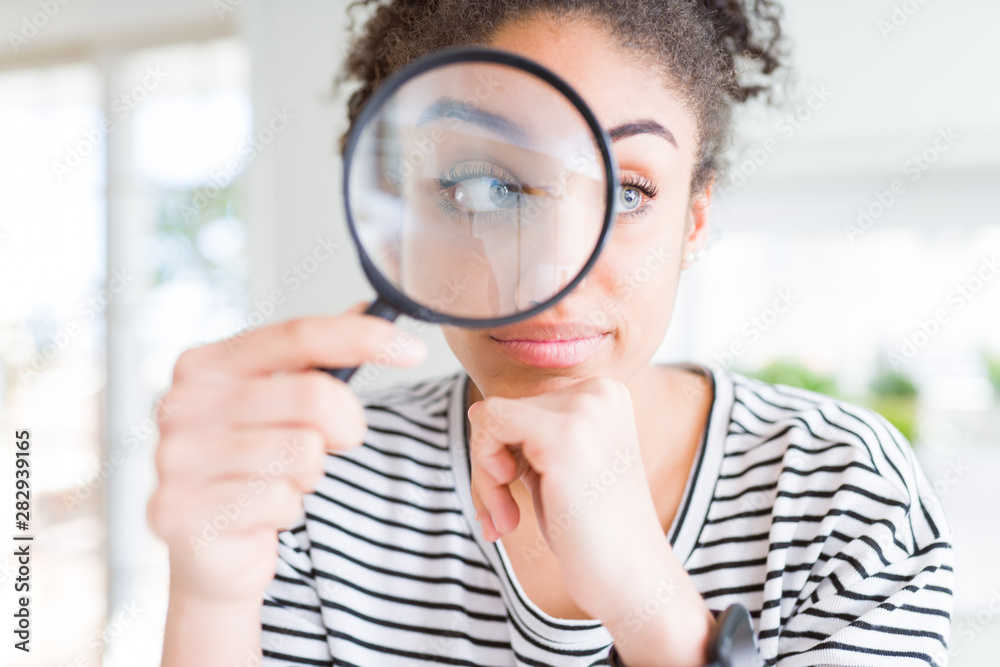 Young african american woman looking through magnifying glass serious face thinking about question, very confused idea Young african american woman looking through magnifying glass serious face thinking about question, very confused idea