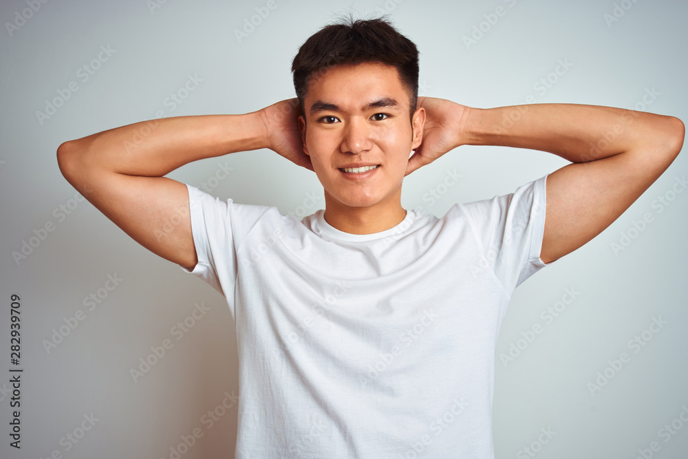 Young asian chinese man wearing t-shirt standing over isolated white background relaxing and stretching, arms and hands behind head and neck smiling happy Young asian chinese man wearing t-shirt standing over isolated white background relaxing and stretching, arms and hands behind head and neck smiling happy