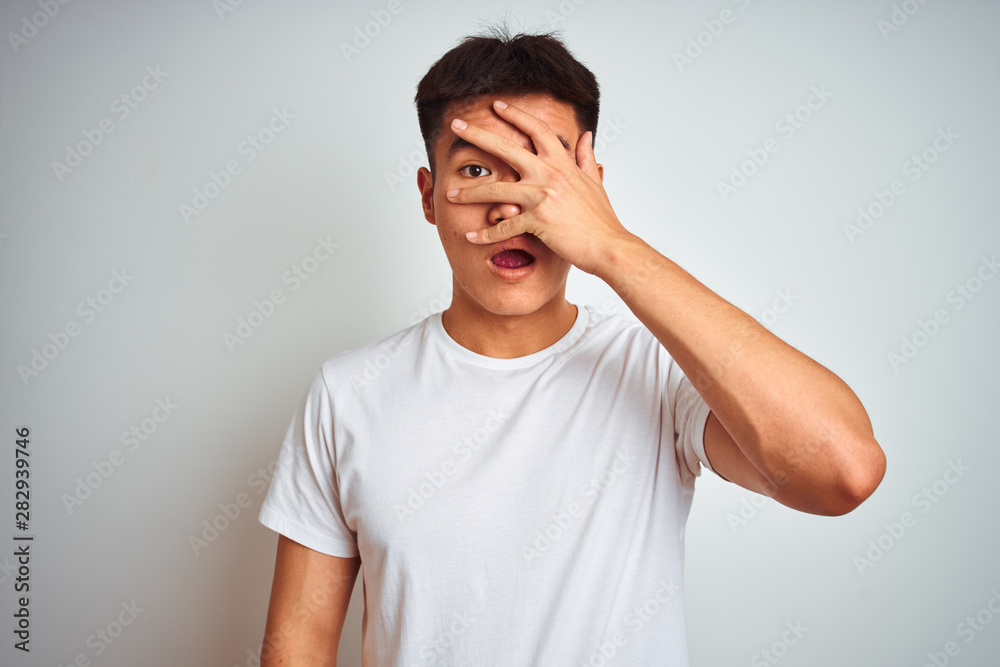 Young asian chinese man wearing t-shirt standing over isolated white background peeking in shock covering face and eyes with hand, looking through fingers with embarrassed expression. Young asian chinese man wearing t-shirt standing over isolated white background peeking in shock covering face and eyes with hand, looking through fingers with embarrassed expression.