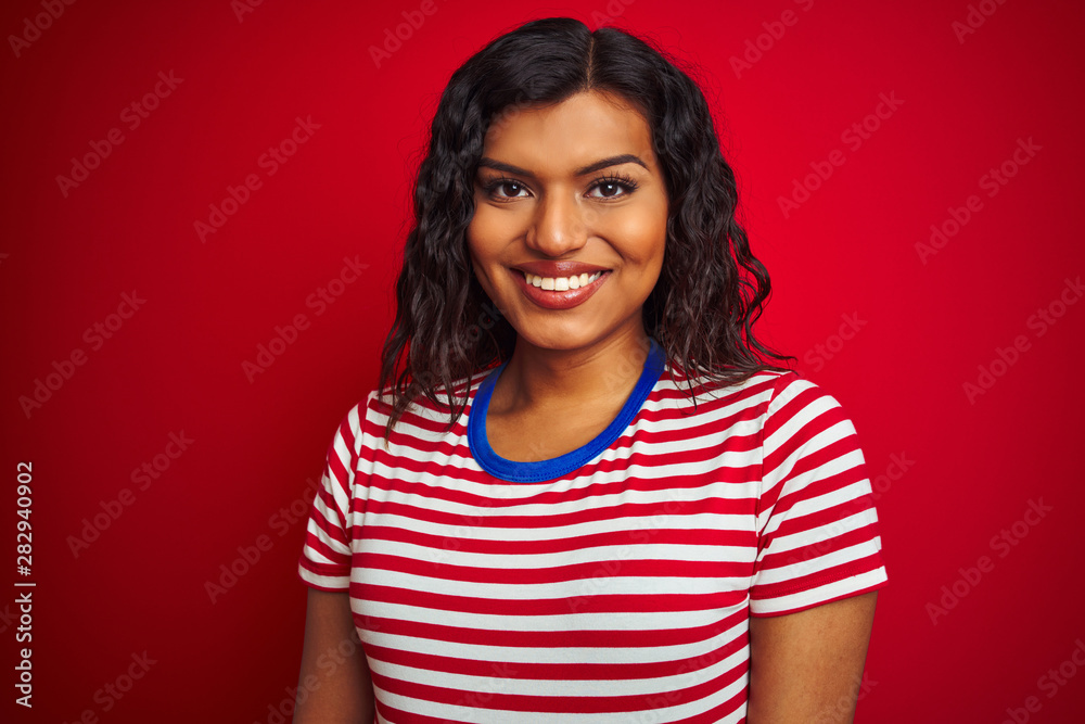 Beautiful transsexual transgender woman wearing stiped t-shirt over isolated red background with a happy face standing and smiling with a confident smile showing teeth Beautiful transsexual transgender woman wearing stiped t-shirt over isolated red background with a happy face standing and smiling with a confident smile showing teeth