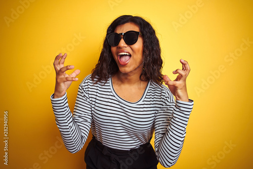 Transsexual transgender woman wearing sunglasses over isolated yellow background crazy and mad shouting and yelling with aggressive expression and arms raised. Frustration concept.