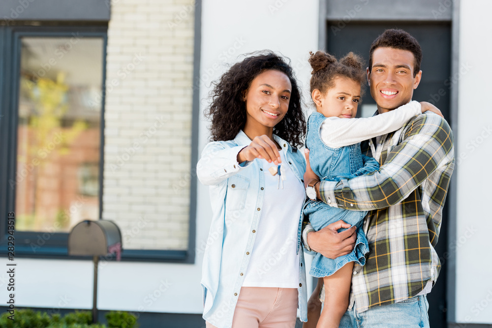 family standing near new house and looking at camera while father holding kid and mother showing key to camera