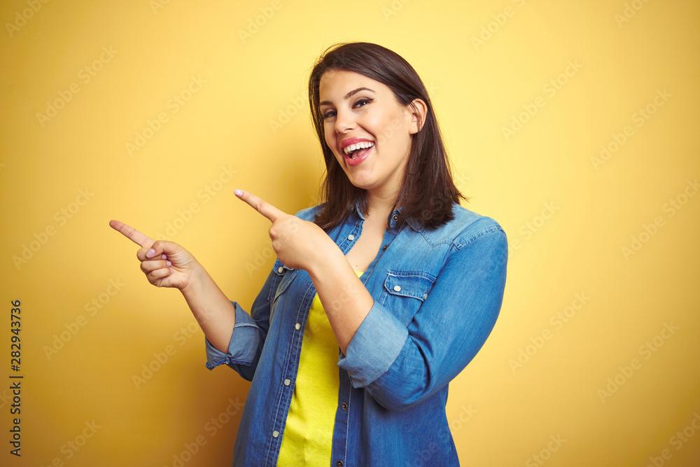 Young beautiful brunette woman wearing casual denim jacket over yellow isolated background smiling and looking at the camera pointing with two hands and fingers to the side. Young beautiful brunette woman wearing casual denim jacket over yellow isolated background smiling and looking at the camera pointing with two hands and fingers to the side.