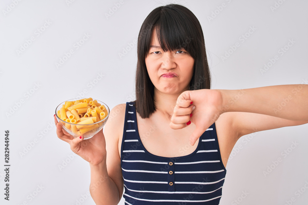 Young beautiful Chinese woman holding bowl with macaroni over isolated white background with angry face, negative sign showing dislike with thumbs down, rejection concept Young beautiful Chinese woman holding bowl with macaroni over isolated white background with angry face, negative sign showing dislike with thumbs down, rejection concept