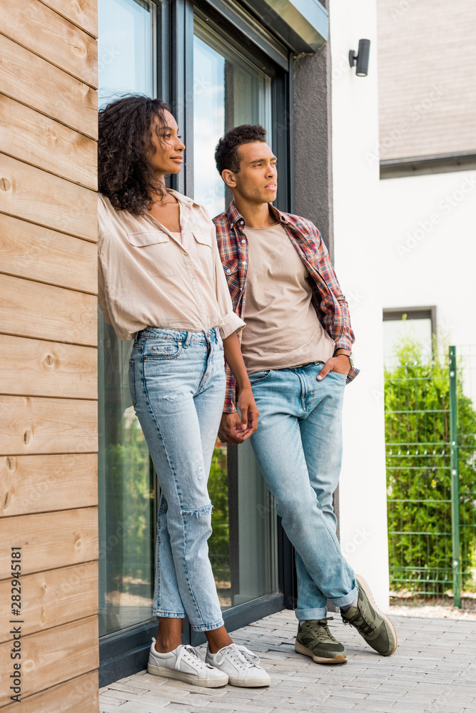 full length view of african american man and woman leaning on door while standing outdoors and looking away