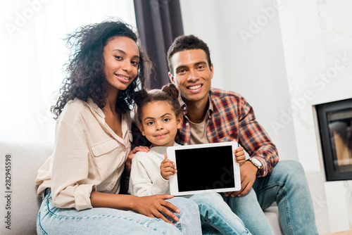kid showing at camera digital tablet while african american parents smiling and looking at camera