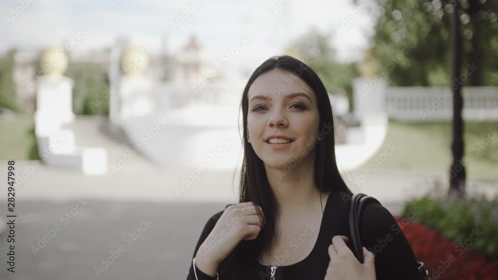 Teenage girl is smiling, looking at the camera and fixing her hair. Portrait of female brunette student in black dress with bag on shoulder in a good mood standing in city park smiles at camera