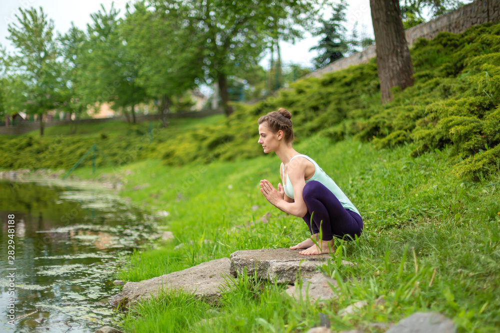 Young beautiful caucasian brunette girl doing yoga on a green lawn against the background of the river