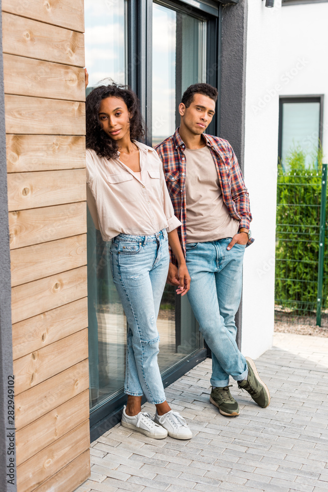 full length view of african american man and woman leaning on door while standing outdoors and looking at camera