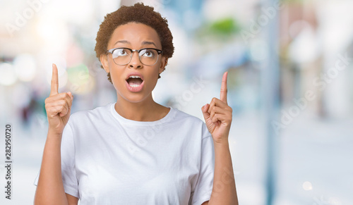 Beautiful young african american woman wearing glasses over isolated background amazed and surprised looking up and pointing with fingers and raised arms.