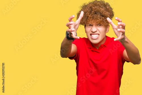 Young handsome man with afro hair wearing red t-shirt Shouting frustrated with rage, hands trying to strangle, yelling mad