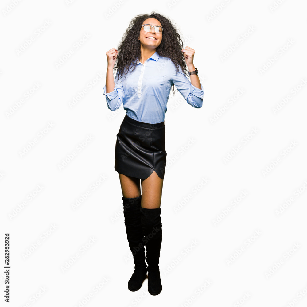 Fototapeta premium Young beautiful business girl with curly hair wearing glasses excited for success with arms raised celebrating victory smiling. Winner concept.