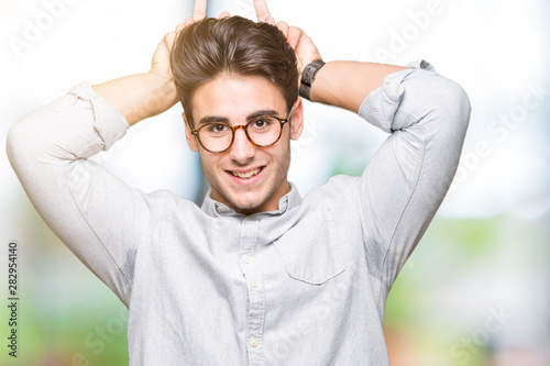 Young handsome man wearing glasses over isolated background Posing funny and crazy with fingers on head as bunny ears, smiling cheerful