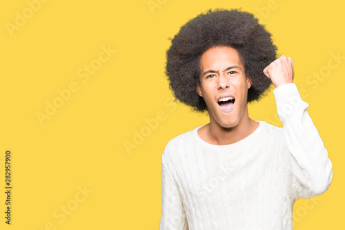 Young african american man with afro hair wearing winter sweater angry and mad raising fist frustrated and furious while shouting with anger. Rage and aggressive concept.