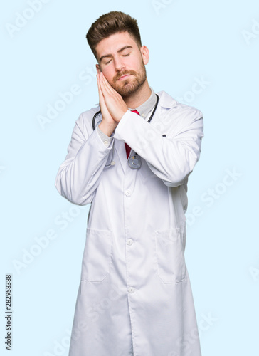 Young handsome doctor man wearing medical coat sleeping tired dreaming and posing with hands together while smiling with closed eyes. © Krakenimages.com