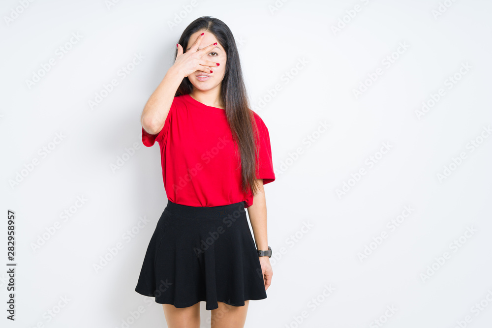 Beautiful brunette woman wearing red t-shirt and skirt over isolated background peeking in shock covering face and eyes with hand, looking through fingers with embarrassed expression. Beautiful brunette woman wearing red t-shirt and skirt over isolated background peeking in shock covering face and eyes with hand, looking through fingers with embarrassed expression.