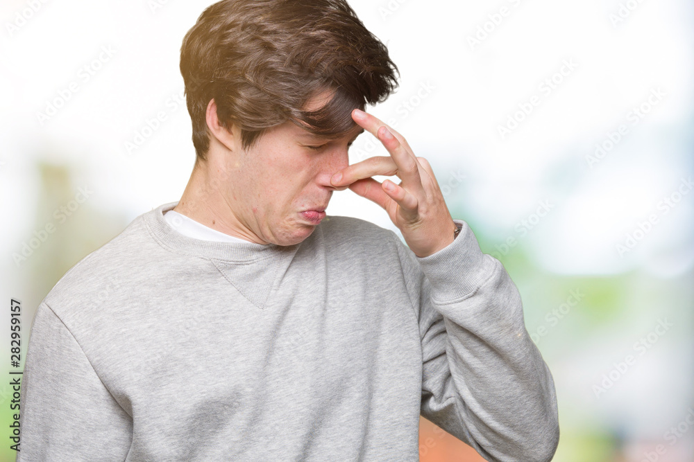 Young handsome sporty man wearing sweatshirt over isolated background smelling something stinky and disgusting, intolerable smell, holding breath with fingers on nose. Bad smells concept.