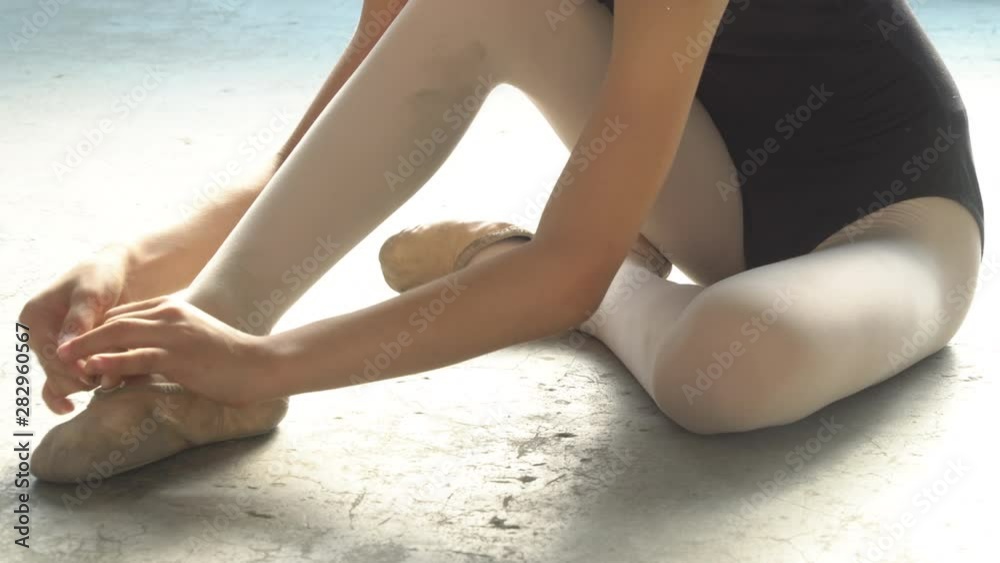 4K Medium close up shot young little child girl ballerina dancer in black leotard for dancing sitting on the floor in studio room wearing and tie ballet slipper shoe for private ballet dance practice. 4K Medium close up shot young little child girl ballerina dancer in black leotard for dancing sitting on the floor in studio room wearing and tie ballet slipper shoe for private ballet dance practice.