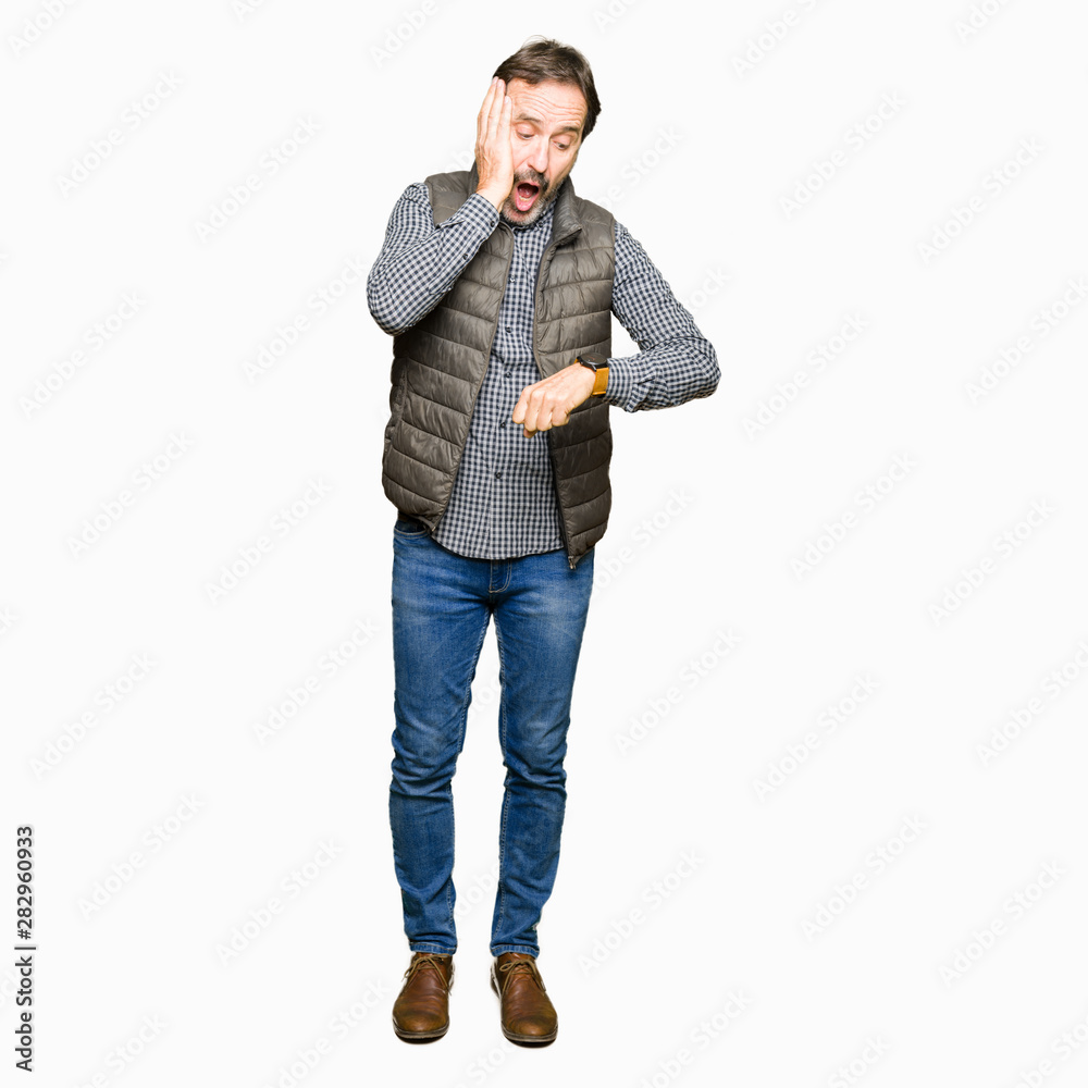 Middle age handsome man wearing winter vest Looking at the watch time worried, afraid of getting late Middle age handsome man wearing winter vest Looking at the watch time worried, afraid of getting late