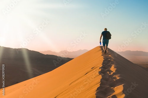A man with backpack goes up on sand dune in sunset. Desert landscape