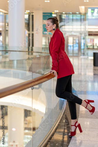 young white girl in red jacket, heels and black trousers leans on railing in mall and looks away. Full height.