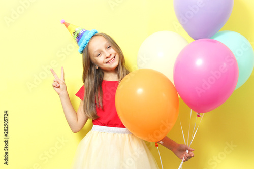 Portrait of a little cute girl in a festive cap and with balloons on a colored background. Birthday, holiday.