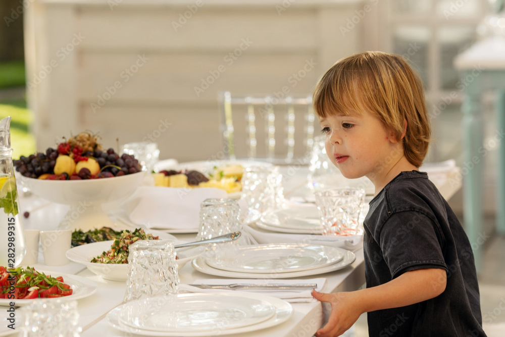 Curious toddler observing festive table exquisitely served for banquet on white tablecloth and waiting for guests Curious toddler observing festive table exquisitely served for banquet on white tablecloth and waiting for guests