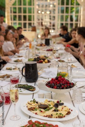 Group of people gathered at long wooden table in restaurant served for party with glasses and cutlery and dishes with fruits and snacks