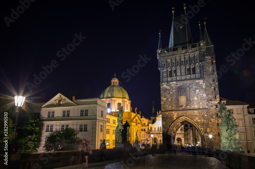 People at the Charles Bridge (Karluv most) and lit St. Francis of Assissi Church and Old Town Bridge Tower at the Old Town in Prague, Czech Republic, in the evening.