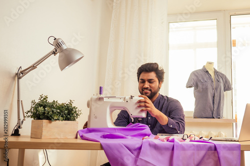 handsome indian tailor man In a stylish shirt workinh with violet cotton textile at home workshop:on the table a lamp and a pot of plants photo