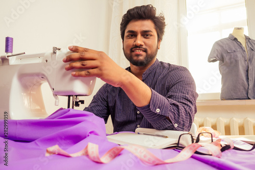 handsome indian tailor man In a stylish shirt workinh with violet cotton textile at home workshop:on the table a lamp and a pot of plants photo