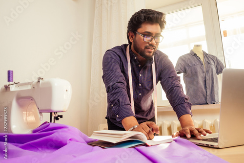 handsome indian tailor man In a stylish shirt workinh with violet cotton textile at home workshop:on the table a lamp and a pot of plants photo