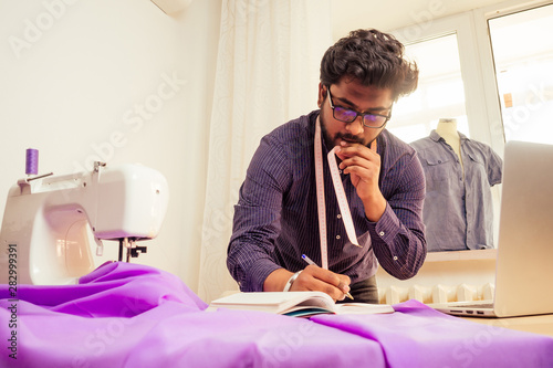 handsome indian tailor man In a stylish shirt workinh with violet cotton textile at home workshop:on the table a lamp and a pot of plants photo