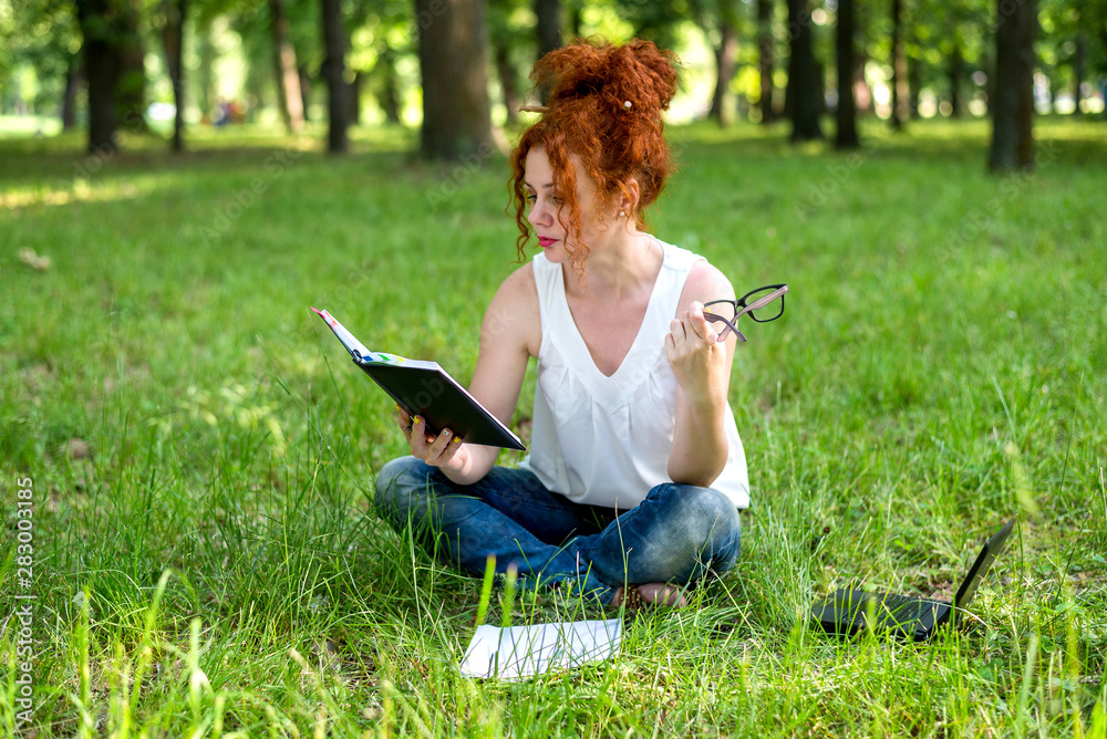 Woman sitting on grass at park working on laptop. Red-haired female freelancer working outdoors
