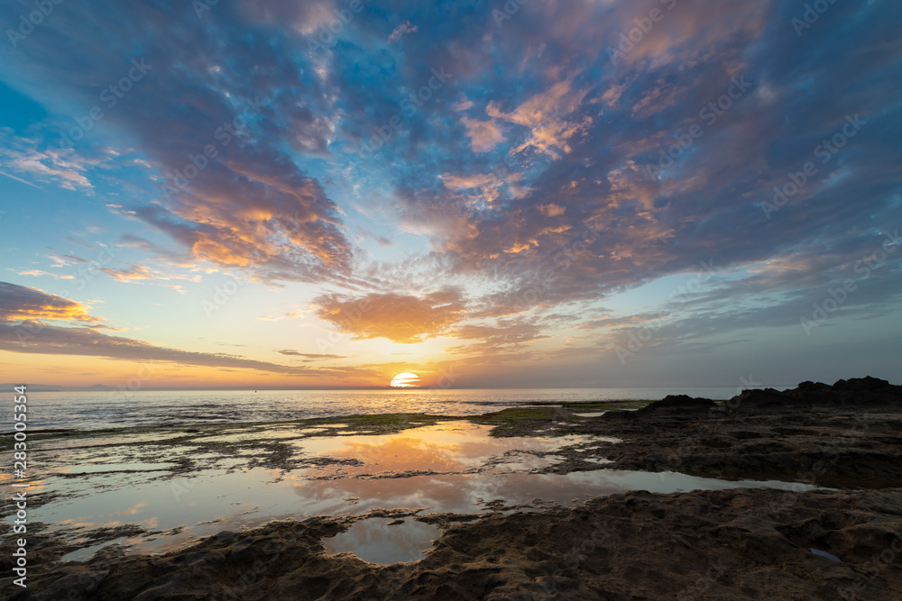 Beautiful sunrise over the Mediterranean sea on the rocky coast of La Mata near the Spanish town of Torrevieja. Colorful clouds adorn the sky with reflections in the water.
