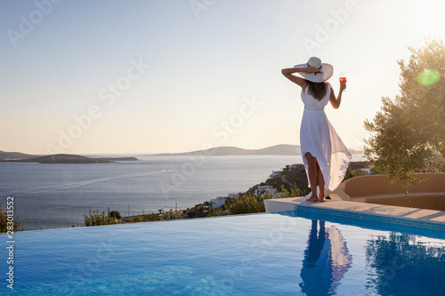 Frau mit weißem Hut steht am Pool und genießt die Aussicht auf das Meer bei Sonnenuntergang mit einem Aperitif in der Hand photo