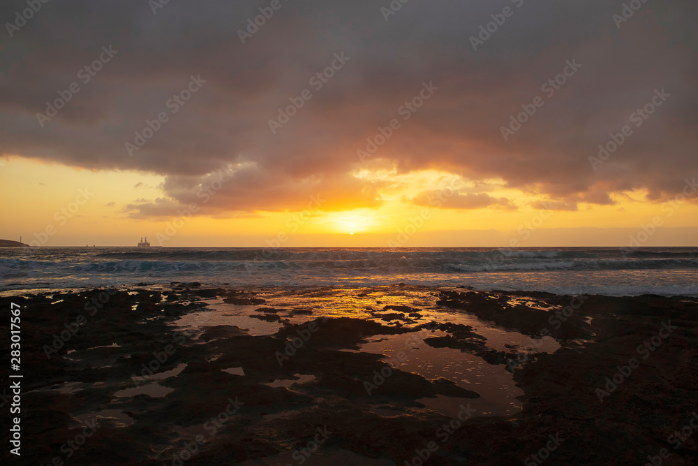 Vibrant, golden sunrise over the limestone coasts of El Medano, Tenerife, Canary Islands, Spain. Mellow light sunrise over the Atlantic with calm waters and a silhouetted oil rig in the distance.