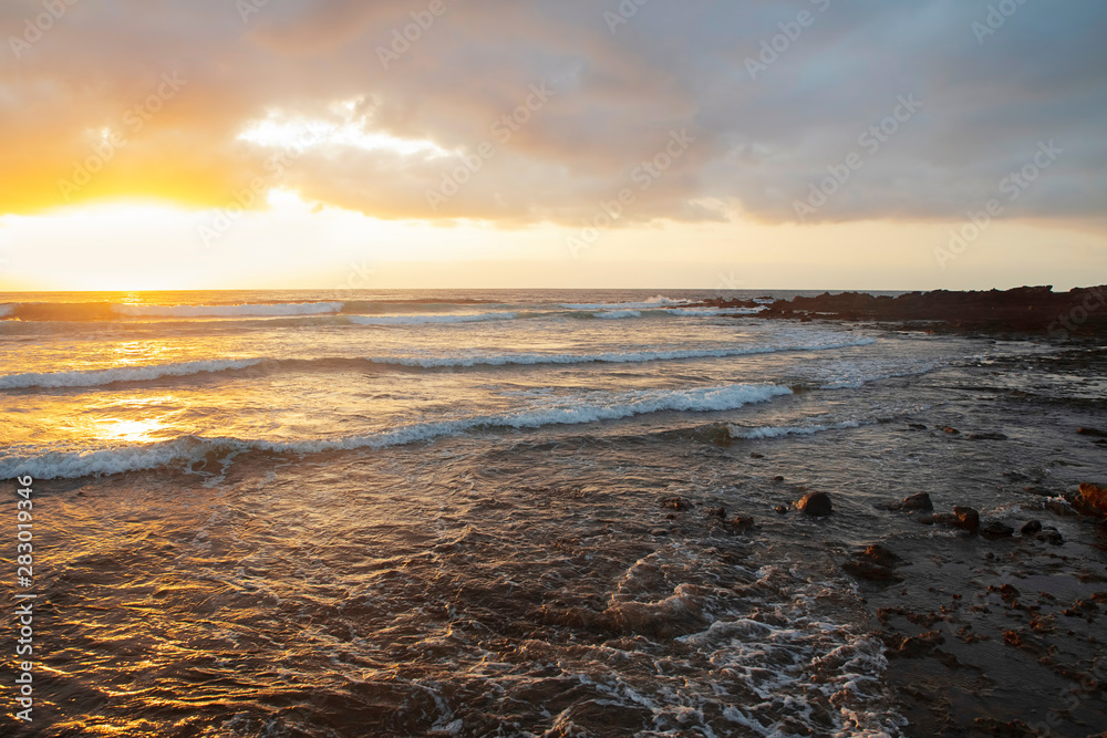 Tropical sunrise over the limestone coasts of El Medano, Tenerife, Canary Islands, Spain. Beautiful seascape with vibrant colors overcast sky, golden haze light and dynamic waves striking rocky shores