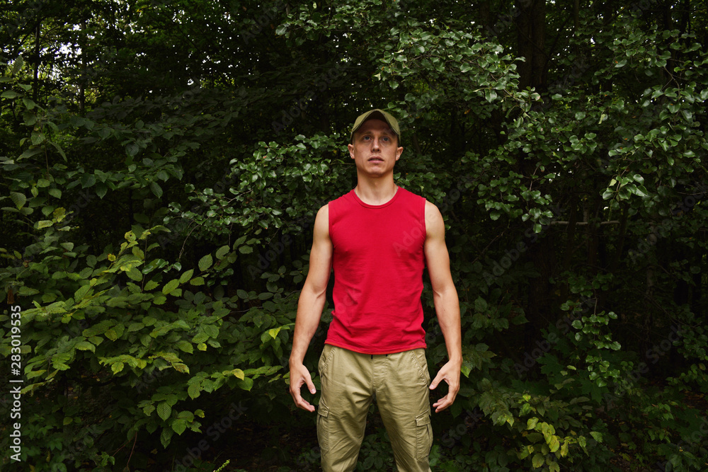 A young man in a red t-shirt, cap and pants on a background of green foliage in the forest.